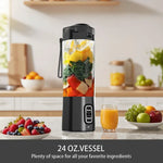 Portable electric fruit mixer 24oz vessel filled with mango, kale, and vegetables on kitchen counter with fresh fruit bowl and glass of orange juice; "24 oz vessel — plenty of space for all your favorite ingredients" text