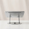 Silver metal laptop stand with 360-degree swivel round base and ventilated tray, rear view showing adjustable folding arms, on beige background